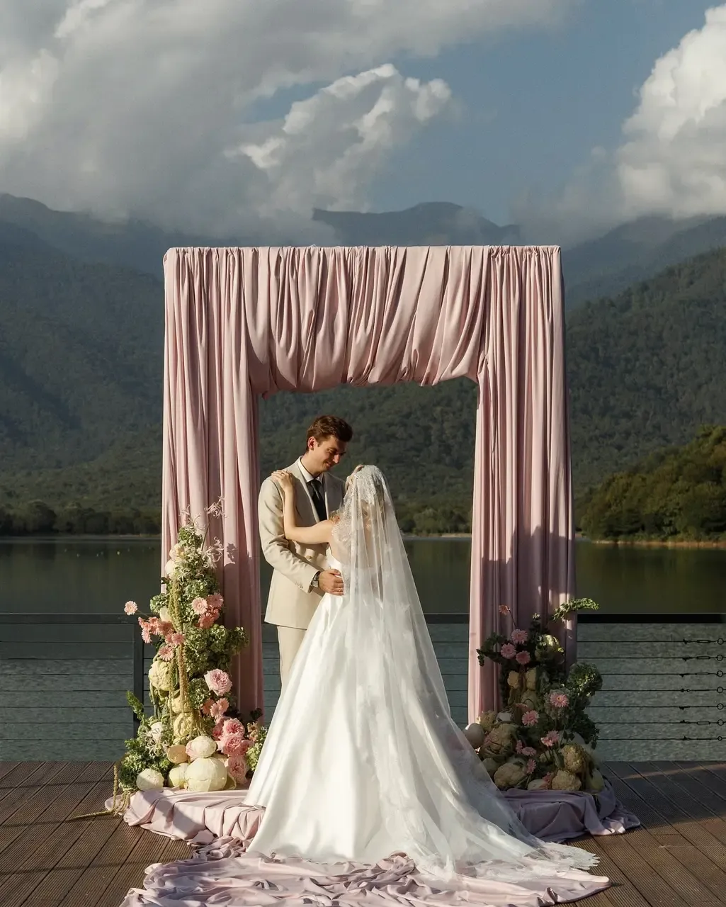 A draped pink fabric arch with floor arrangements of roses on a lakeside deck, decor by WEDDING DEKOR