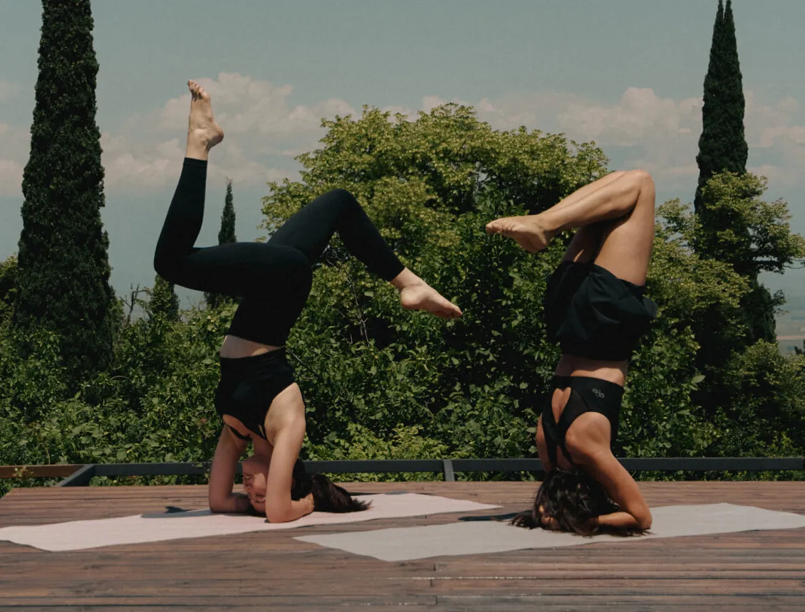 Two women in headstands on the wooden yoga terrace of Horizons Bakurtsikhe among cypress trees