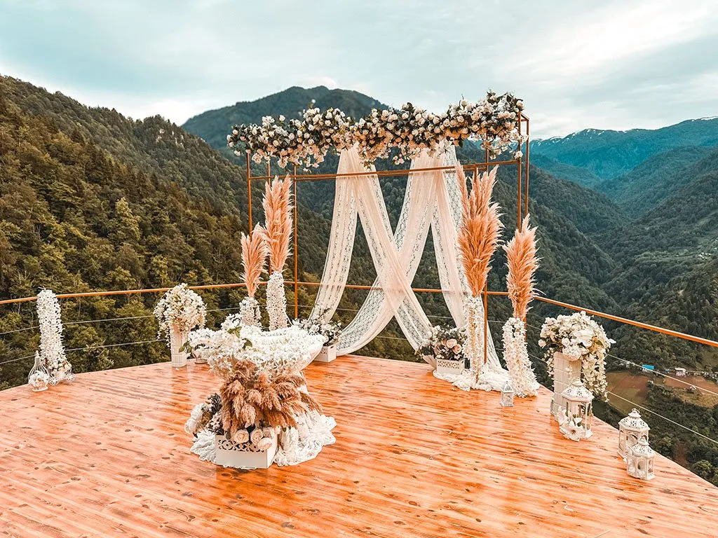 A ceremony arch with white flowers and pampas grass on a wooden platform above a forested valley