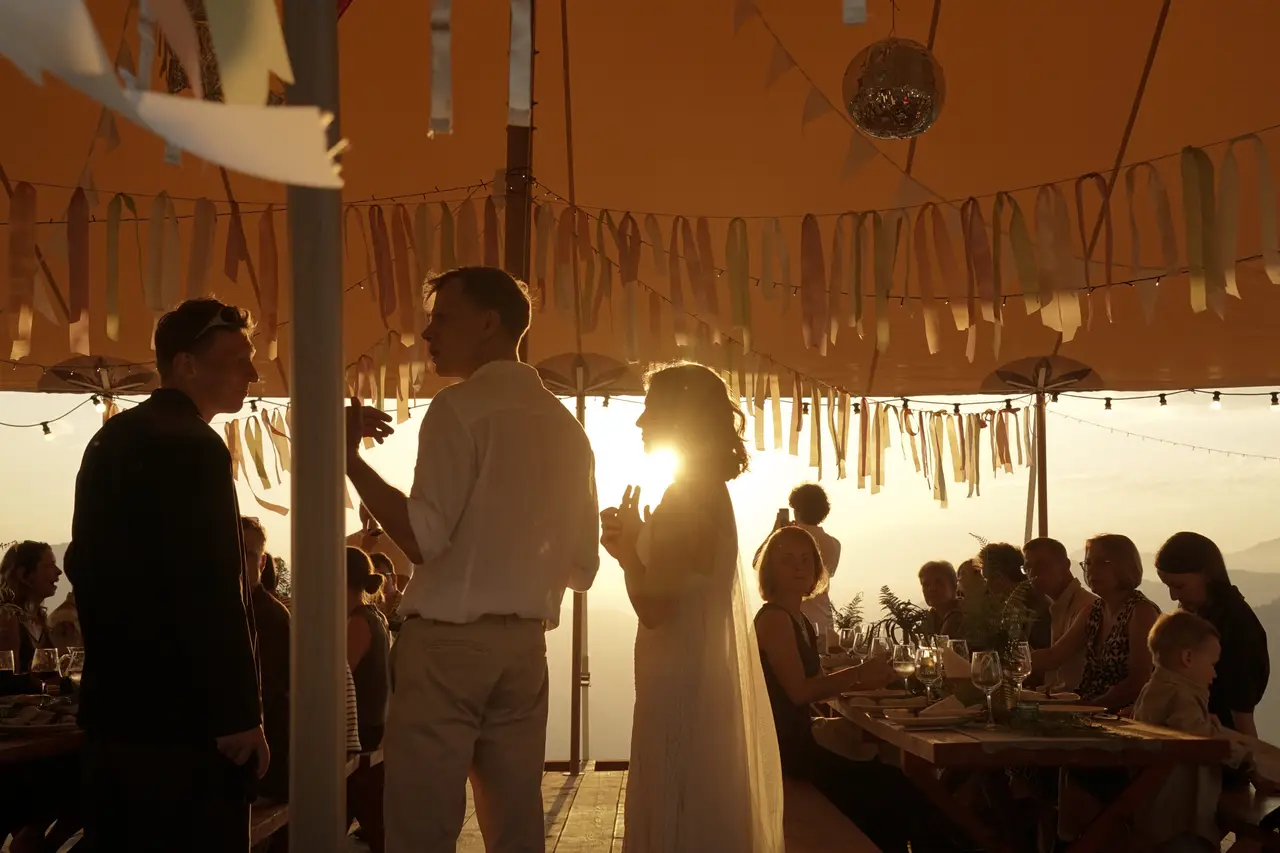 Guests in silhouette under an event tent hung with ribbons and string lights at sunset