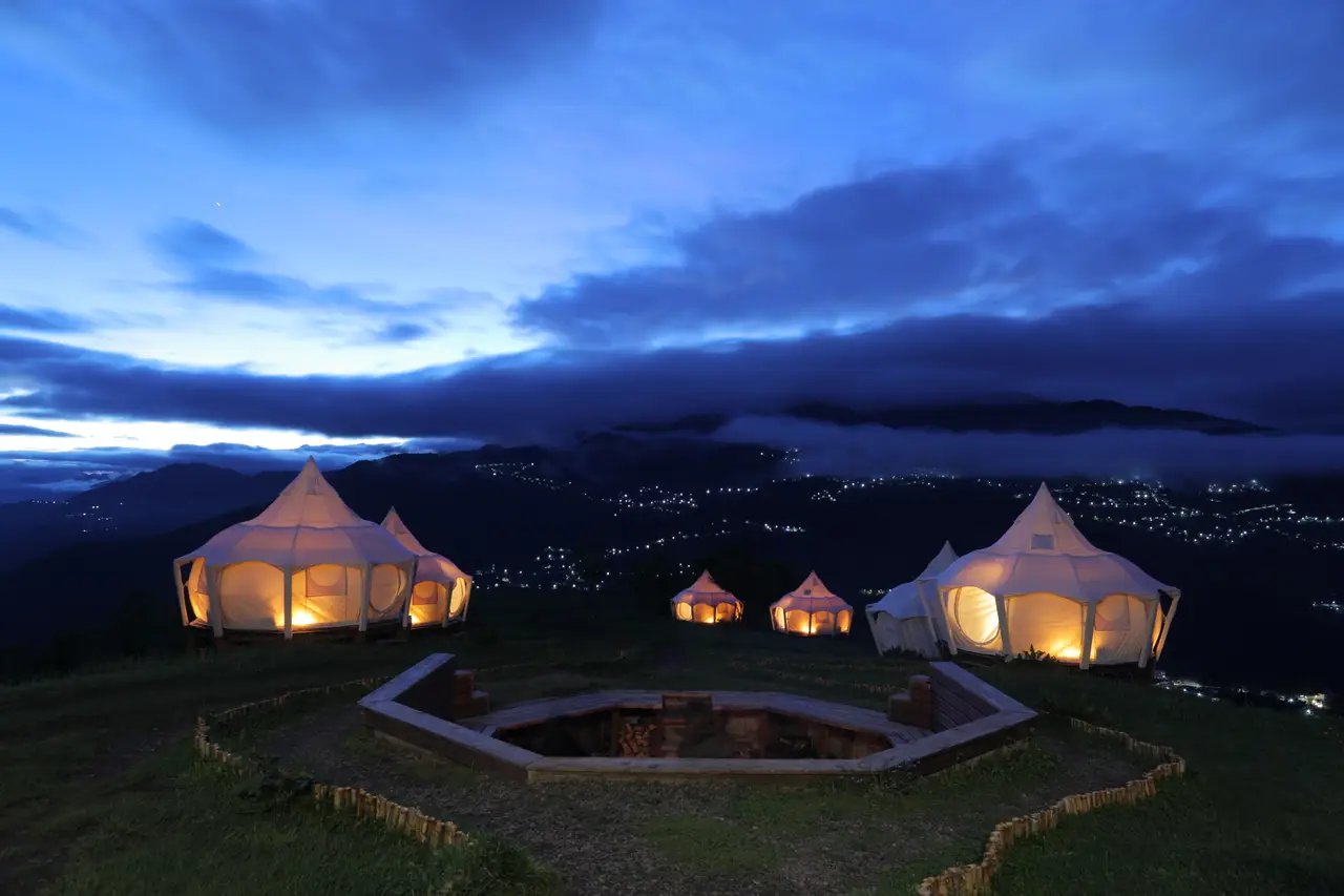 განათებული კარვები კოცონის წრის გარშემო Glamping Tago-ში საღამოს, ქვემოთ სოფლის შუქებით