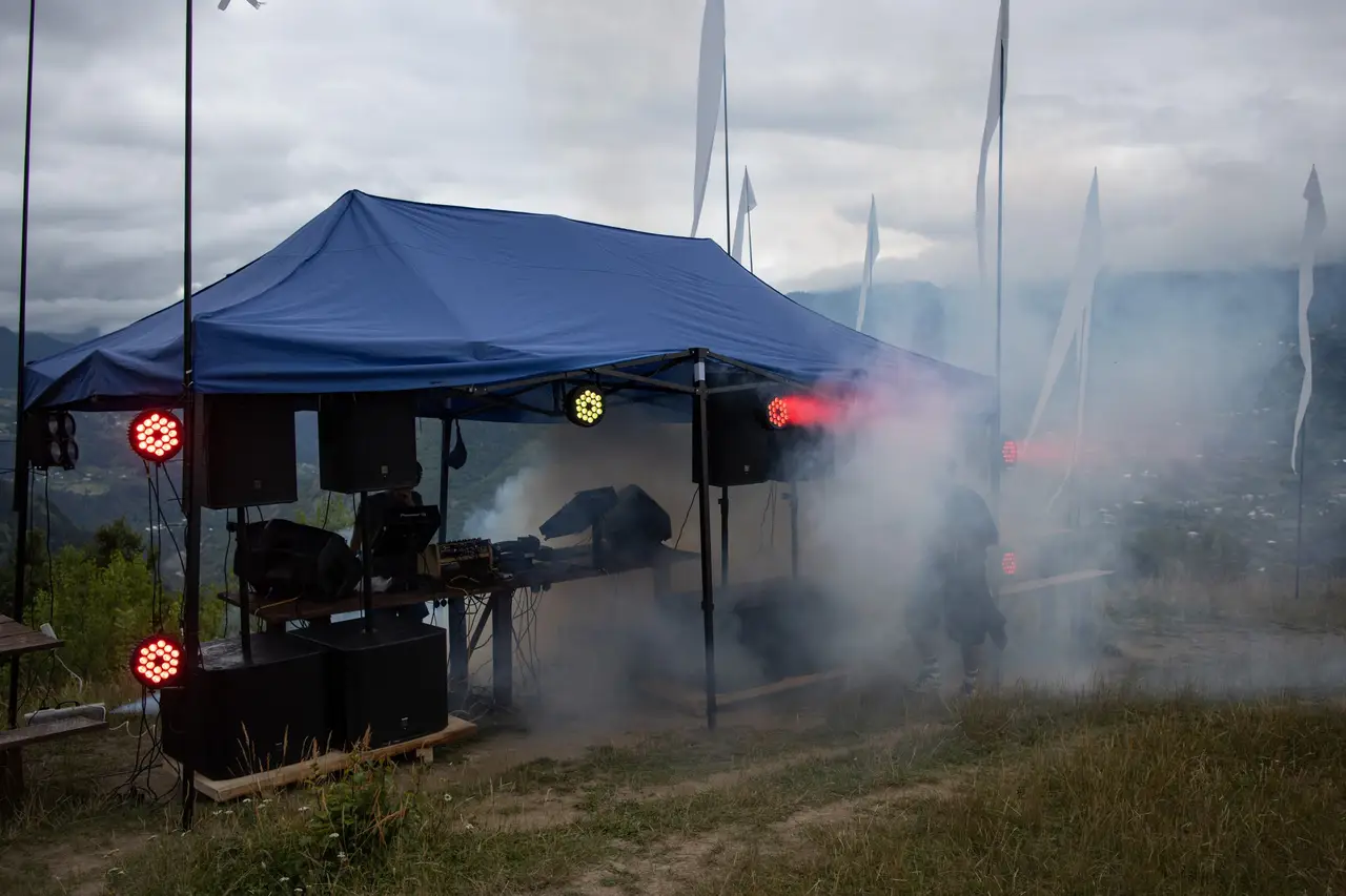 A DJ set-up with coloured lights and smoke under an event tent on a mountain hillside