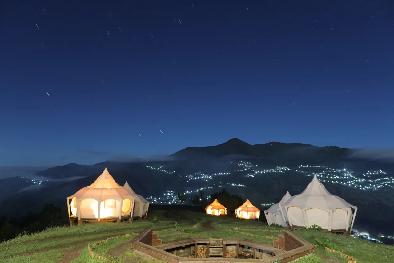 Glamping Tago tents lit up at night under a starry sky, a campfire pit in front