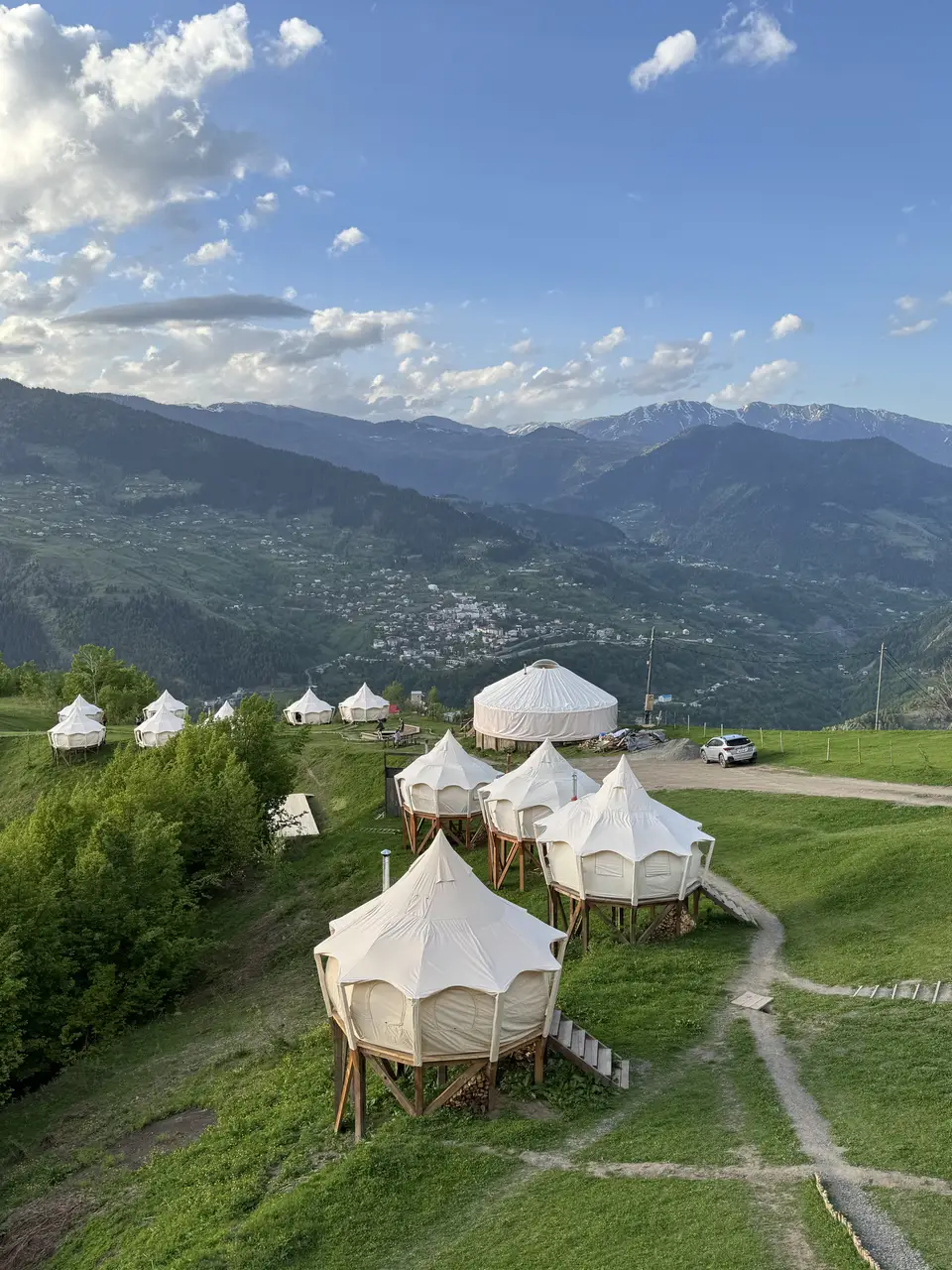 Aerial view of the white tents of Glamping Tago on a green ridge above a valley with villages and distant snowy mountains