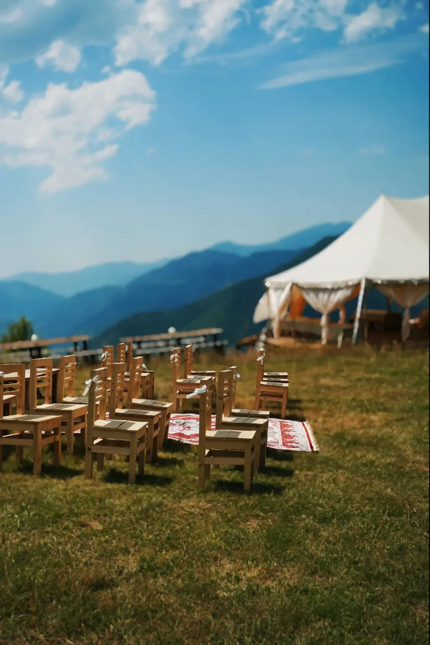Wooden chairs with white ribbons on the grass for an outdoor ceremony, with the event tent and mountains behind